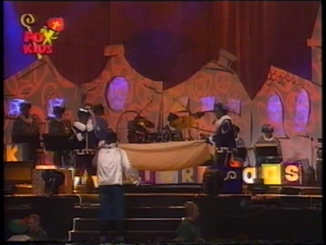 Wegwijspiet (links), Hoofdpiet (rechts) en Muziekpiet (midden) met op de achtergrond het orkest o.l.v. John van Eijck